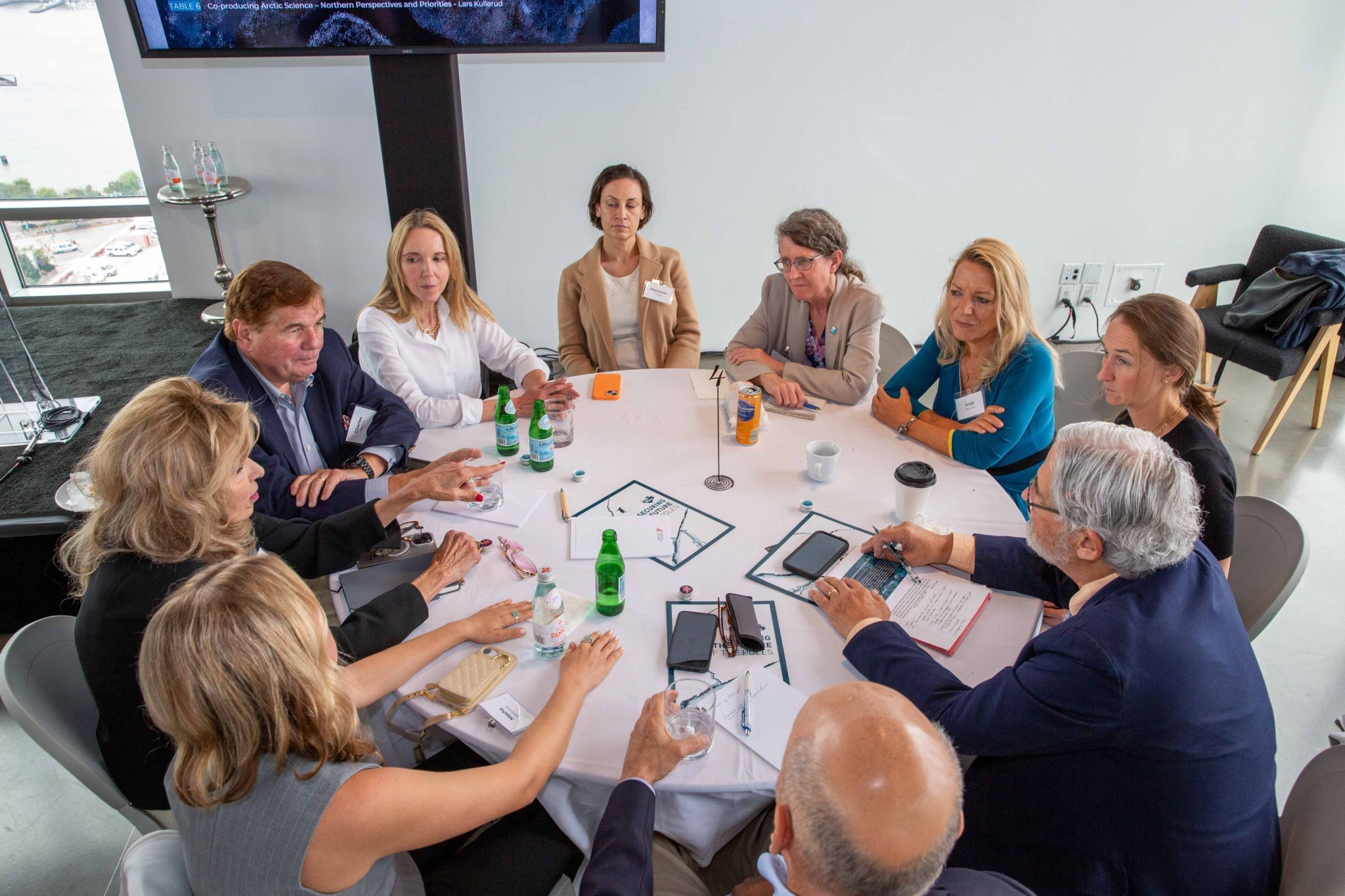 Ten people in professional attire sit around a round table discussing climate science and policy. In the background is a television monitor and a white wall.