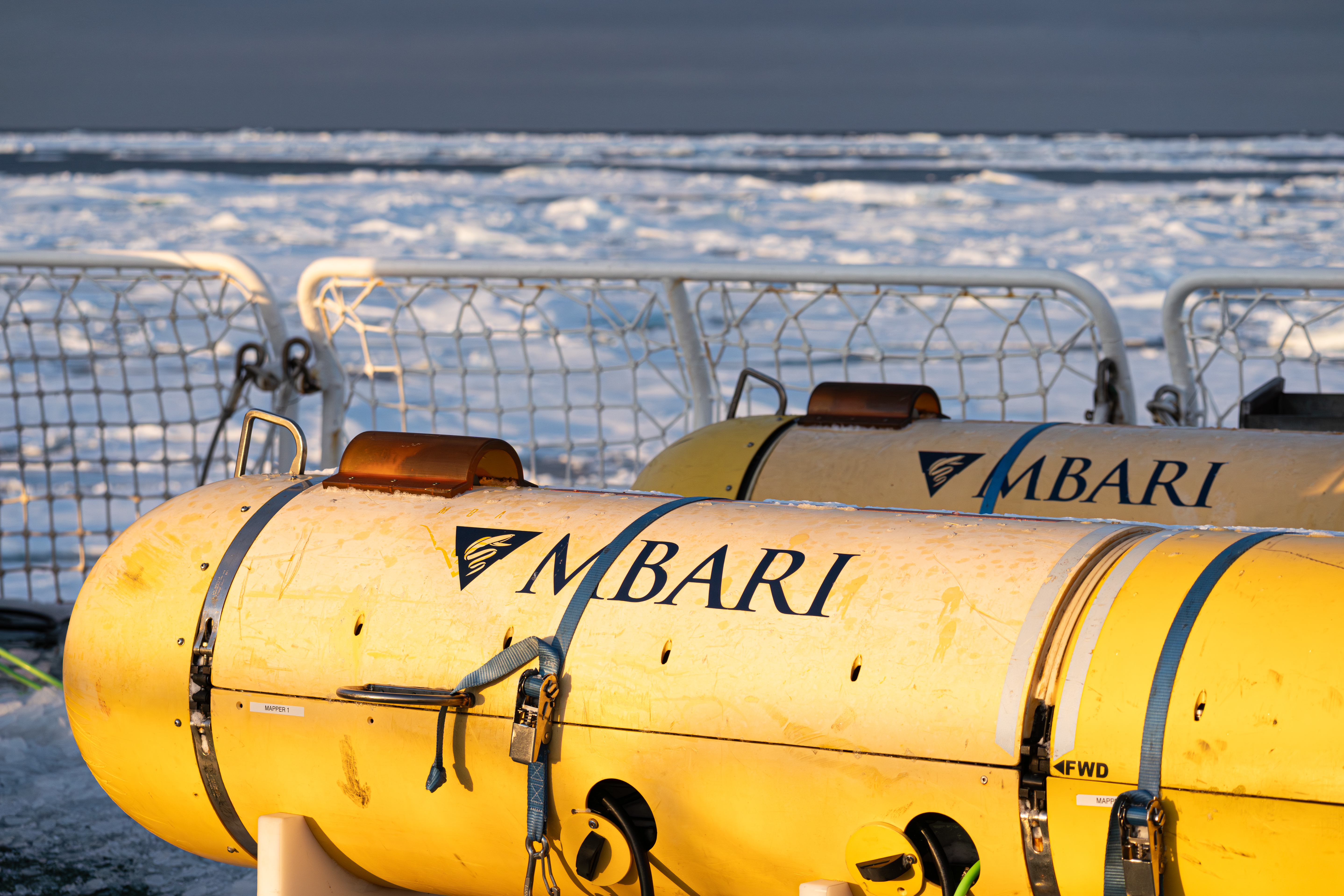 Two seafloor mapping robots sit on the deck of a research vessel. The robots have a cylindrical yellow plastic housing printed with the MBARI logo. The robots are secured with nylon straps. In the background is the ship’s white metal and mesh railing with white ice and gray sky on the horizon.