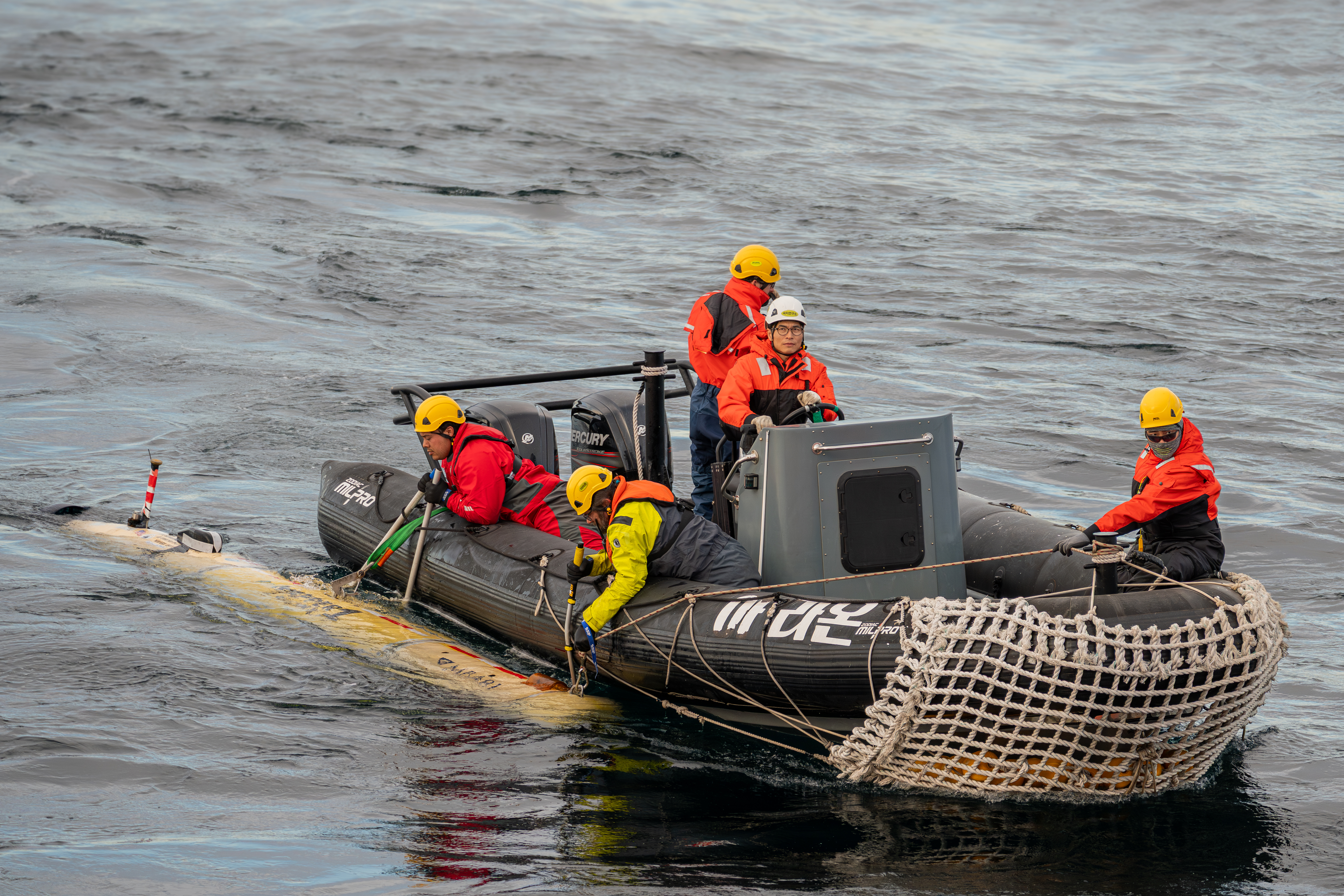 Five marine operations crew retrieve a seafloor mapping robot aboard an inflatable black Zodiac boat. The crew are wearing hard hats, bright orange life vests, and bright orange jackets. Two crew members are securing a cylindrical robot with a yellow plastic housing. The background is gray ocean.