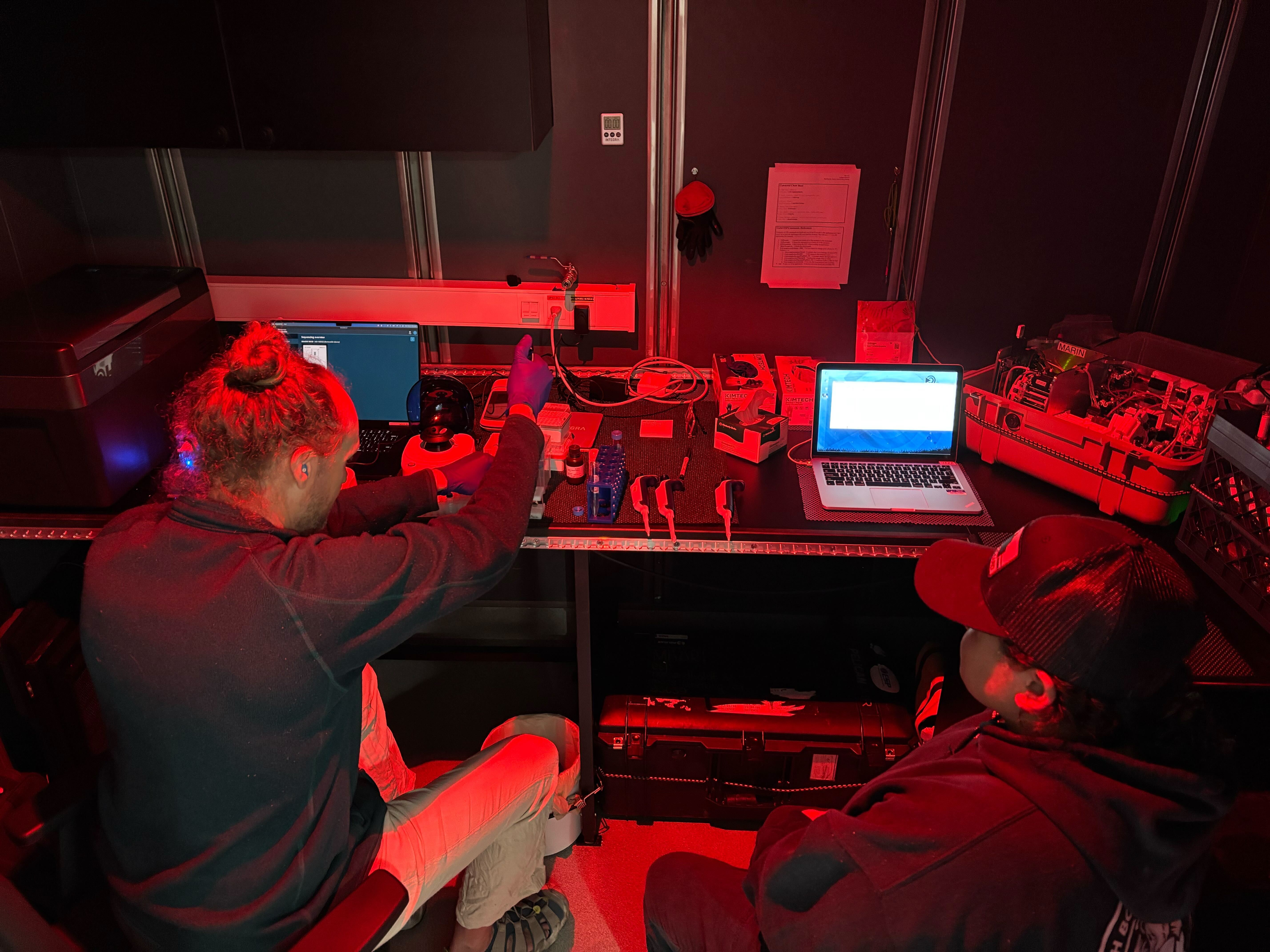 Two researchers conduct DNA sequencing in a lab. The researcher on the left has blond hair in a bun and is wearing a dark jacket and beige pants. The researcher on the right is wearing a black cap, a black hooded sweatshirt, and dark pants. They are seated in front of a black countertop with two laptop computers, several pipettes, and other assorted scientific instruments. The background is the black wall of a shipboard science lab. The frame is illuminated in red light.