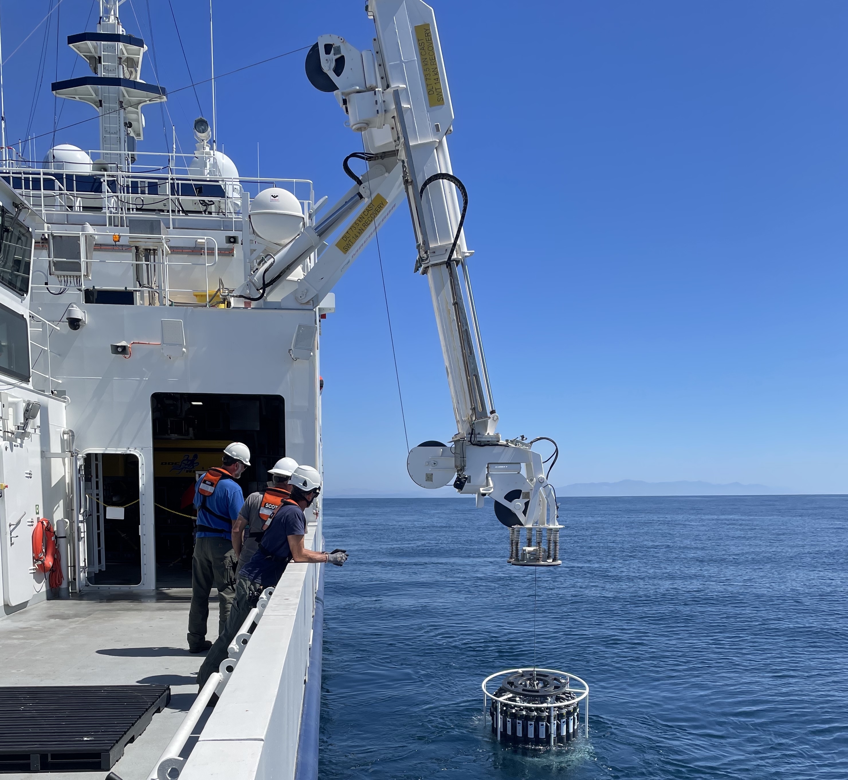 Marine operations crew deploy a scientific instrument over the side of a research ship. On the left side of the frame, three crew members wearing white hard hats, orange life vests, blue shirts, and dark pants stand on the gray metal deck of a ship, leaning against a white metal railing. On the right side of the frame, a white metal crane lowers a round scientific instrument with a carousel of gray plastic bottles. In the background are still blue ocean and blue sky, with hazy clouds and coastal mountains on the horizon.