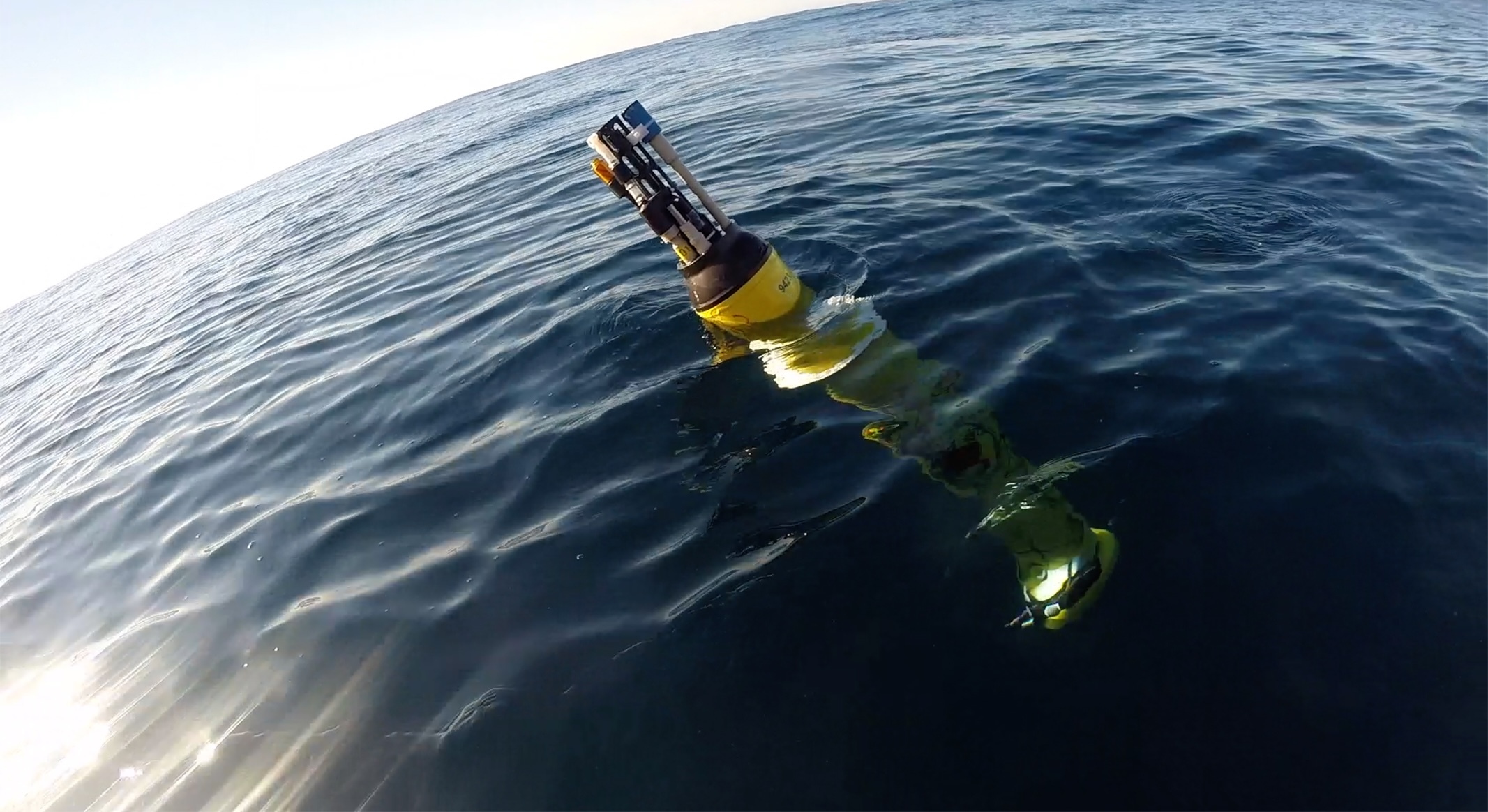 A robotic float drifts at the ocean surface on a sunny day. The cylindrical float has a yellow plastic housing. At the top are white and black sensors protruding out of the water. The background is blue ocean with the bright reflection of the sun in the lower left corner.