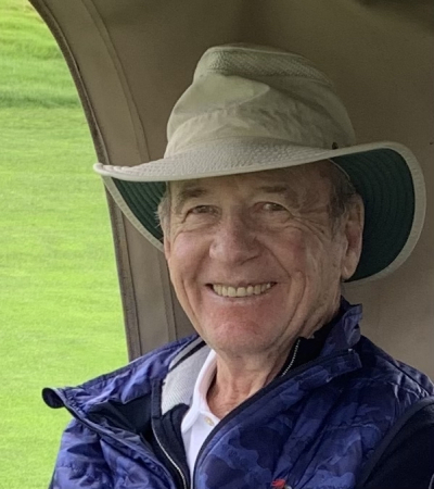 A headshot shows a former member of the MBARI Board of Directors wearing a tan bucket hat, white polo shirt, black quarter-zip sweater, and purple vest. The background is a tan cloth tent and bright-green grass.
