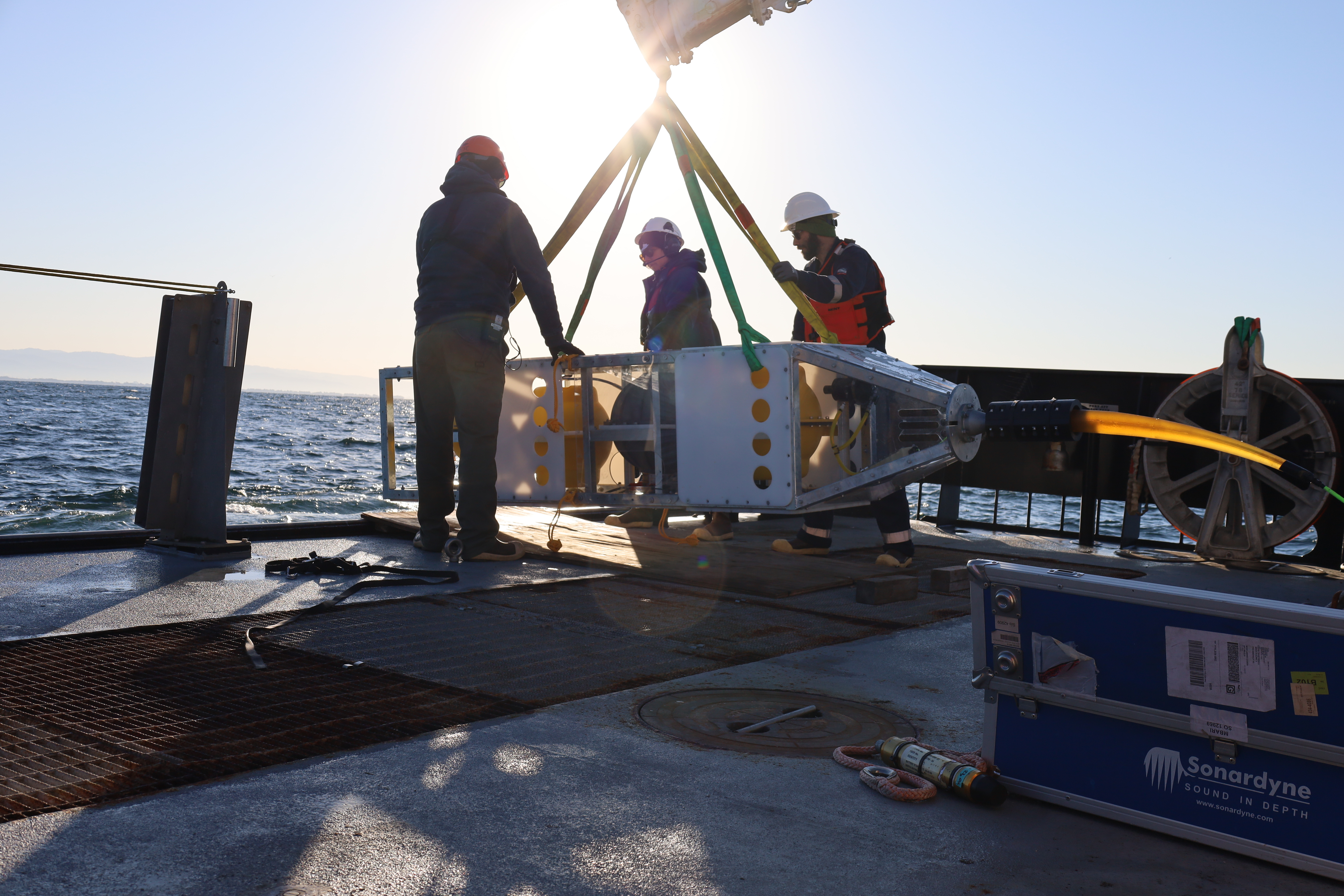 Three MBARI staff prepare for deployment of a scientific instrument on the gray metal deck of a research vessel. The researcher on the left is wearing an orange hard hat, a black hooded sweatshirt, and dark pants. The deckhand in the middle is wearing a white hard hat, sunglasses, and a dark hooded jacket. The engineer on the right is wearing a white hard hat, sunglasses, a navy-blue jacket, an orange life vest, navy-blue pants, and brown boots. All three are in silhouette in front of the bright sun. The scientific instrument has a rectangular silver metal frame, white plastic panels, a silver metal sphere, and a yellow plastic sphere, and is being moved with three yellow nylon straps and one green nylon strap attached to a crane out of frame. In the background is blue ocean, with coastal mountains and blue sky in the distance on the horizon.