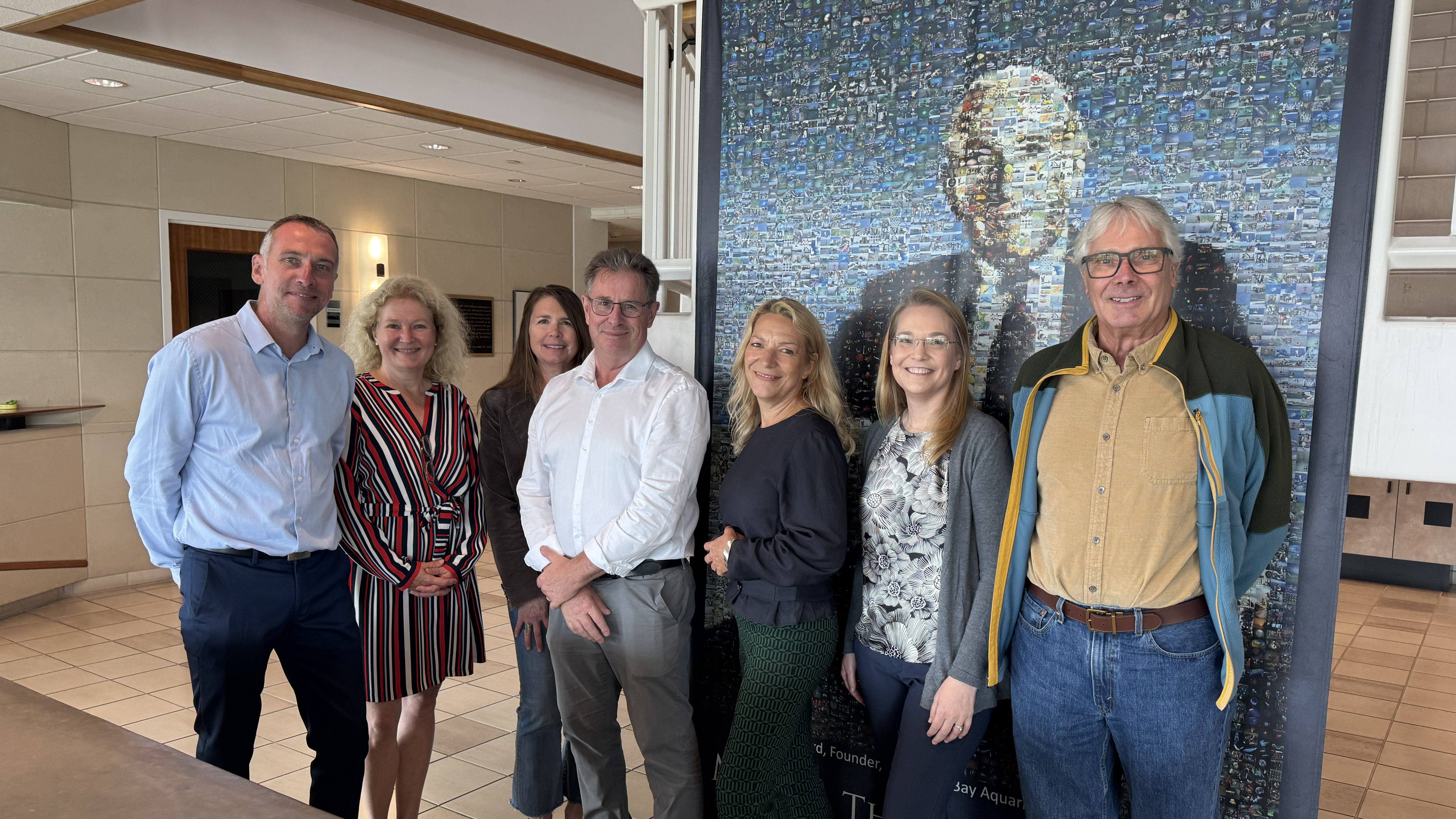 A group of seven people stands in front of a blue photomosaic portrait of MBARI’s founder. In the background are the off-white beige walls, cabinetry, and tile floor of an office building.