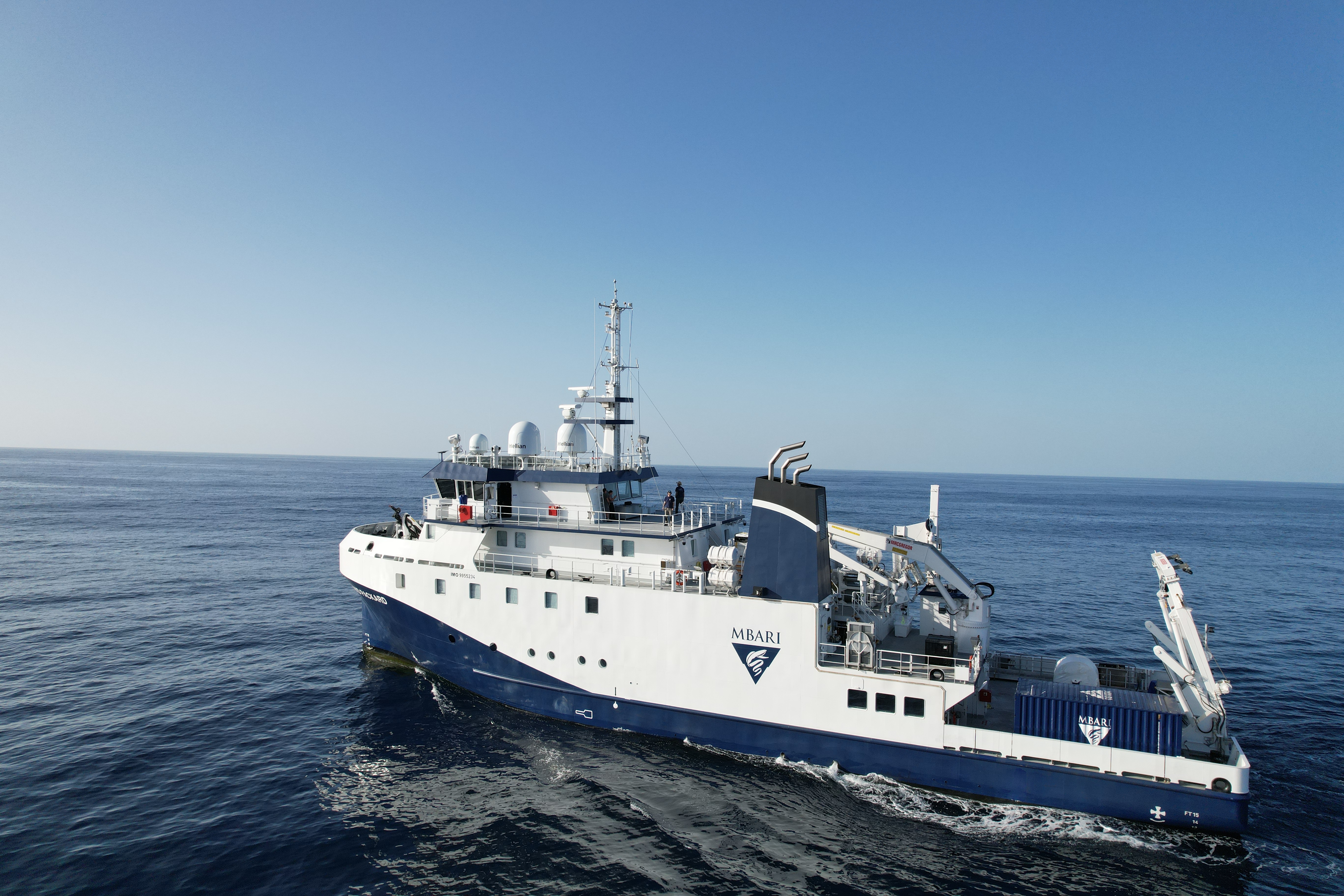 A research ship with a blue hull and white sides sails across the open ocean. The background is still blue water with clear blue sky on the horizon.