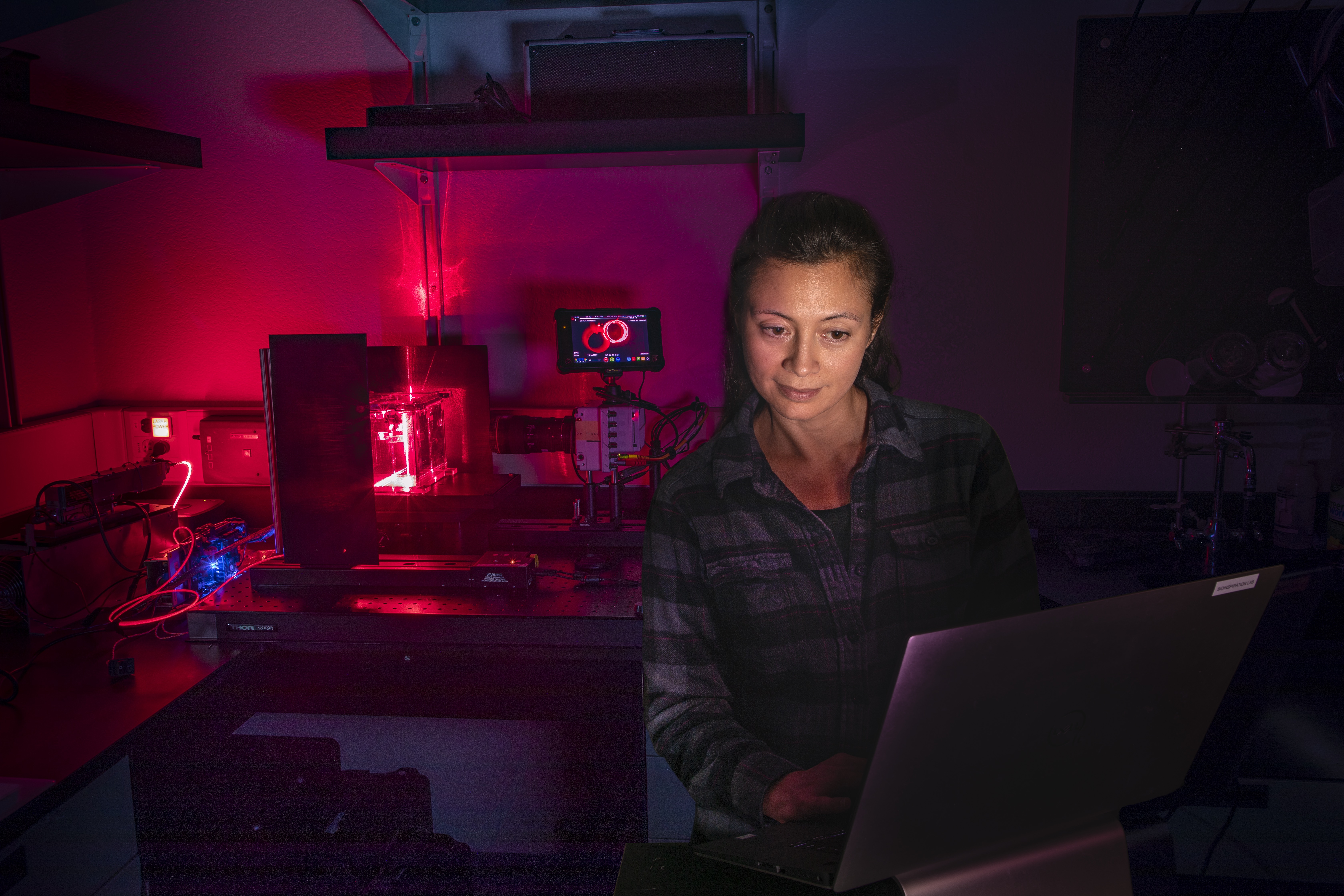 Alt text: A woman with dark hair wearing a dark colored shirt looks at a laptop on a desk in front of her. She is facing the photographer, and the monitor’s light shines in her face, making it the brightest part of the scene. The rest of the scene is very dark with red light in the back behind her, to the left of the frame. The red light gives the background a red glow. There are numerous instruments behind her on a countertop.