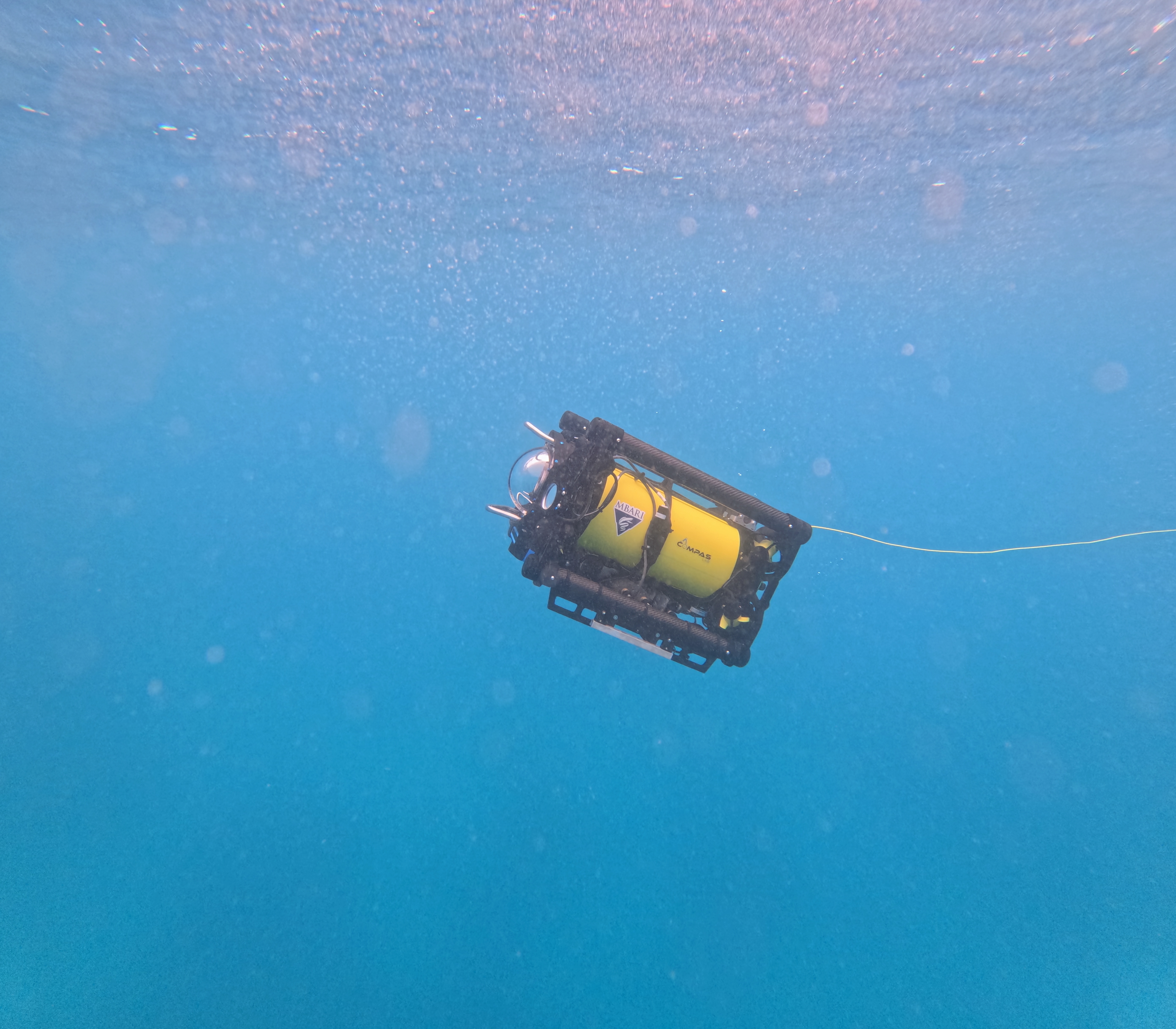A robot moves through open water. The robot has a black metal frame outside with a clear glass dome at the front, facing the left side of the frame. Inside is a yellow metal cylinder with stickers for MBARI and the CoMPAS Lab. A thin yellow cable extends from the robot off frame to the right. The background is bright-blue water.