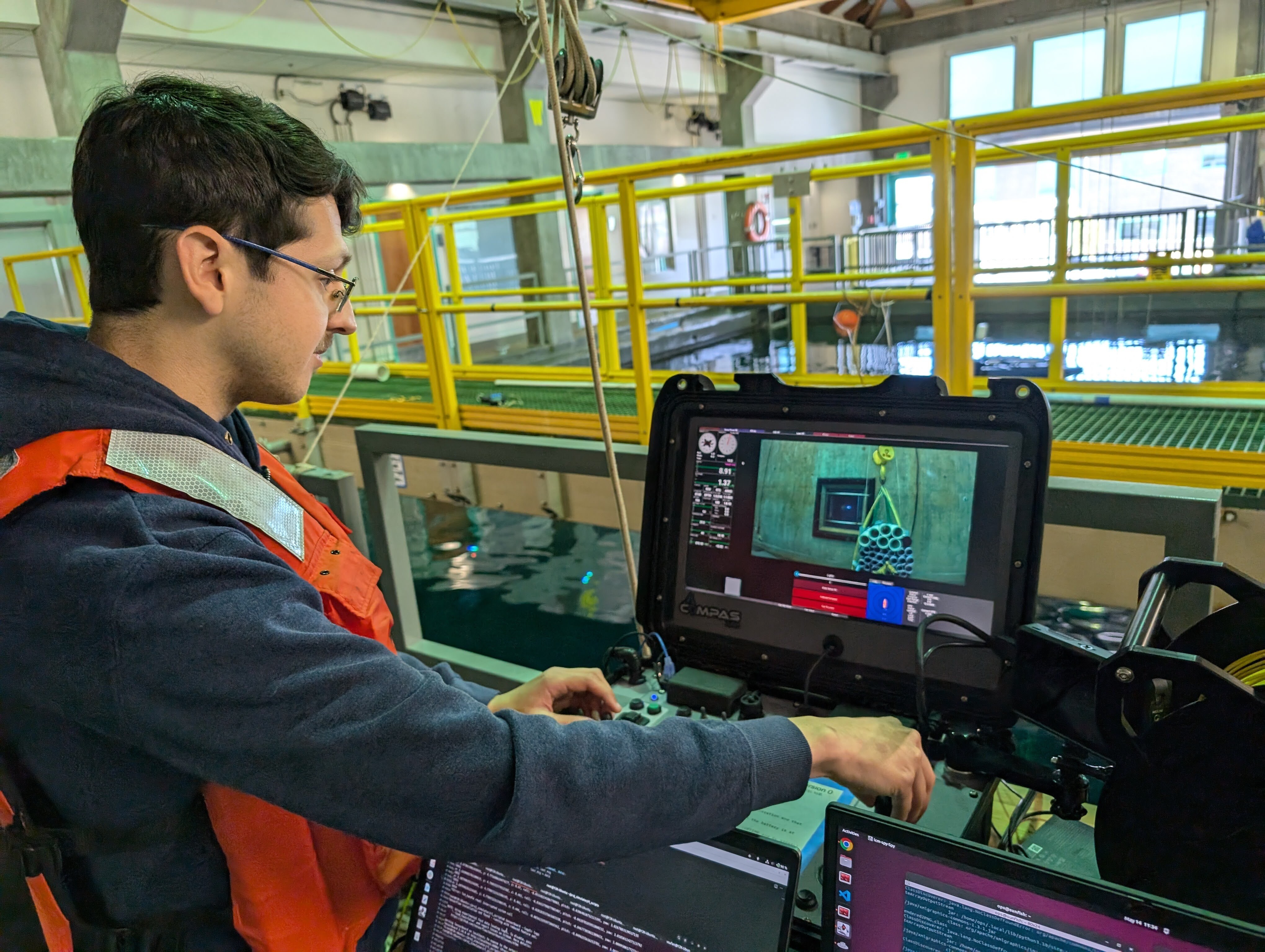 An engineer adjusts the controls for an underwater robot. The engineer has short brown hair and a mustache and is wearing a blue hooded sweatshirt and an orange life vest. He is standing in front of a counter with three computers and watching a live video feed from the robot in the tank below. The engineer is facing a large tank of water with a yellow-and-green fiberglass walkway above and assorted scientific equipment in the background.