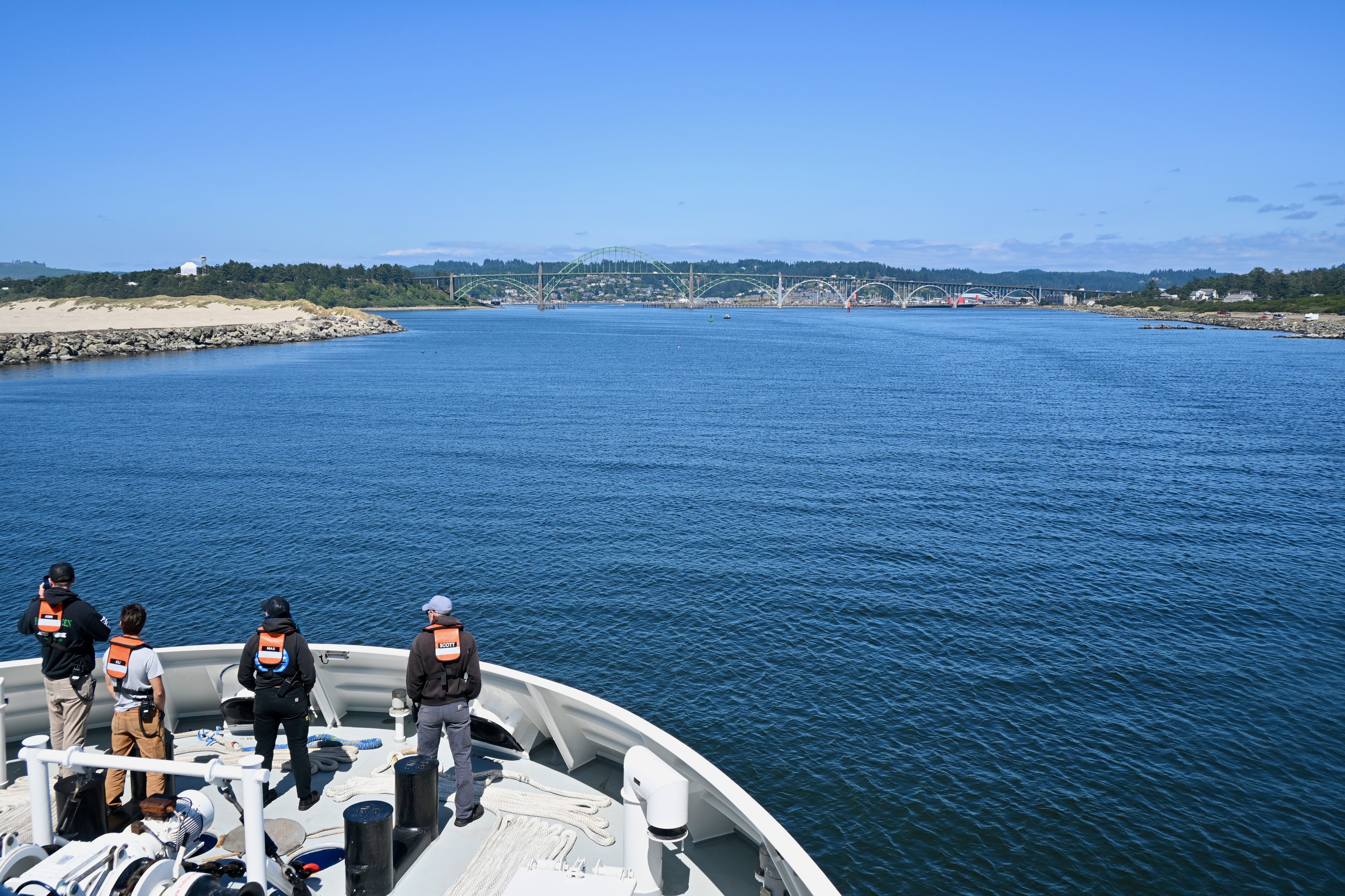 Four members of MBARI’s marine operations crew stand on the gray metal deck of a research vessel during its transit into port. The crew members are all wearing orange life vests. In the background are greenish-blue water, a sandy shoreline, green forests, a gray bridge, and a coastal community, with pale-blue sky overhead.