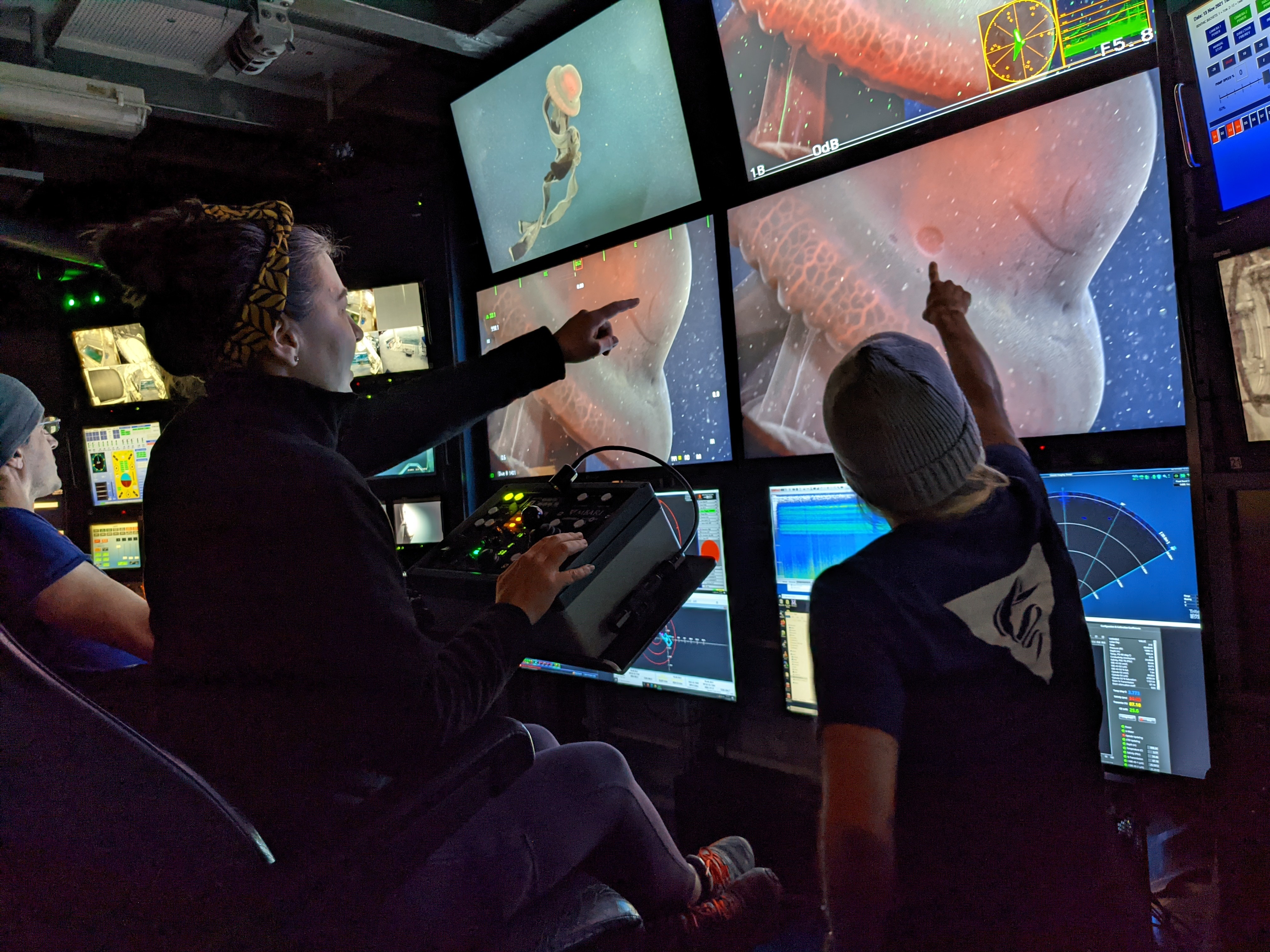 Two researchers in the dark control room aboard a research ship point to a bank of large video monitors displaying a dark red deep-sea jelly. The researcher on the right is wearing a yellow patterned headband and a dark jacket. She is seated with her right hand on a panel of camera controls and her left hand pointing to the video monitor. The second researcher is wearing a gray beanie and a blue t-shirt with the MBARI logo on the back. She is facing away from the camera and pointing to the screen with her right hand.