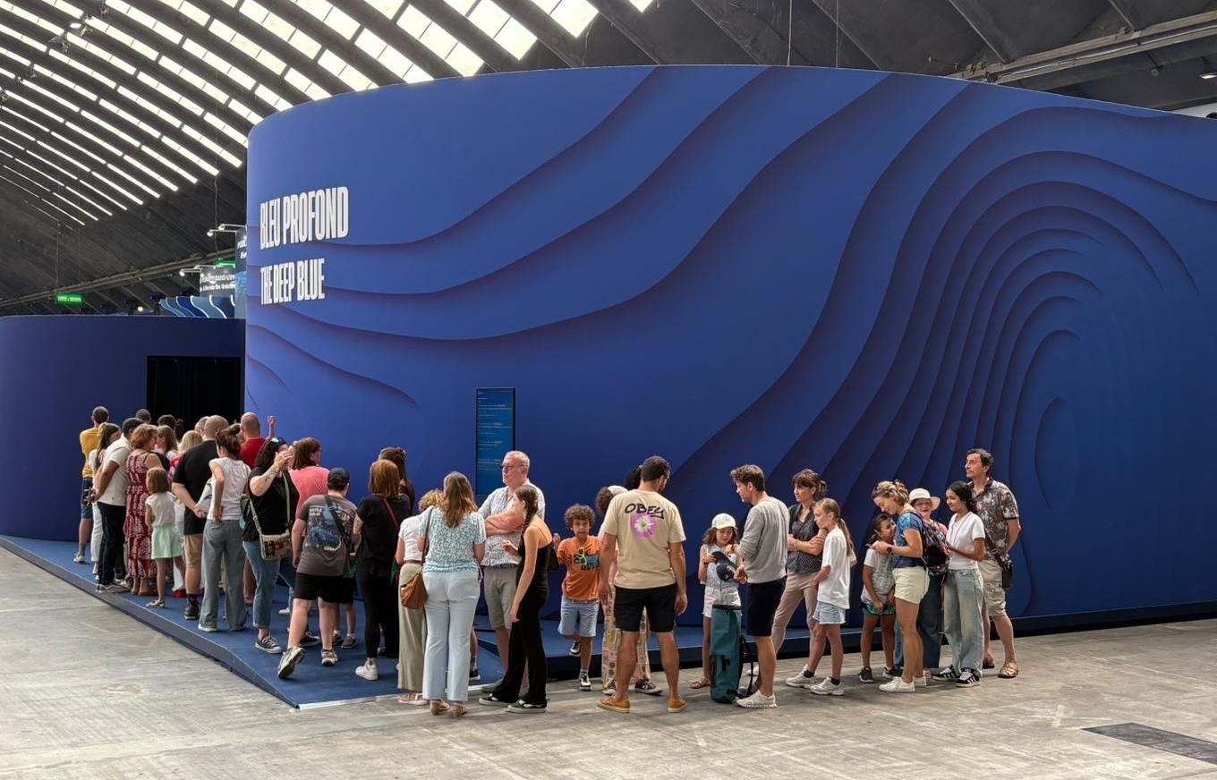 A line of several dozen visitors waits to enter an exhibit hall. The hall is surrounded by rounded, textured walls with large white text reading Bleu Profond The Deep Blue. The foreground is gray floor tile. The background has gray walls with skylights overhead.