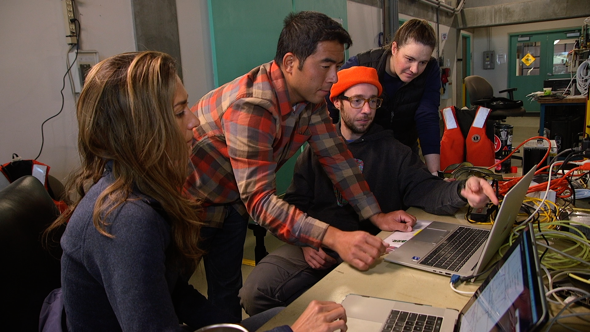 Four MBARI researchers reviewing data on a computer. Two researchers are seated and two are standing. One in the center wearing an orange beanie is pointing to the screen of a silver laptop computer to the right on top of a beige tabletop. Cables and assorted equipment are visible on the table and in the background.