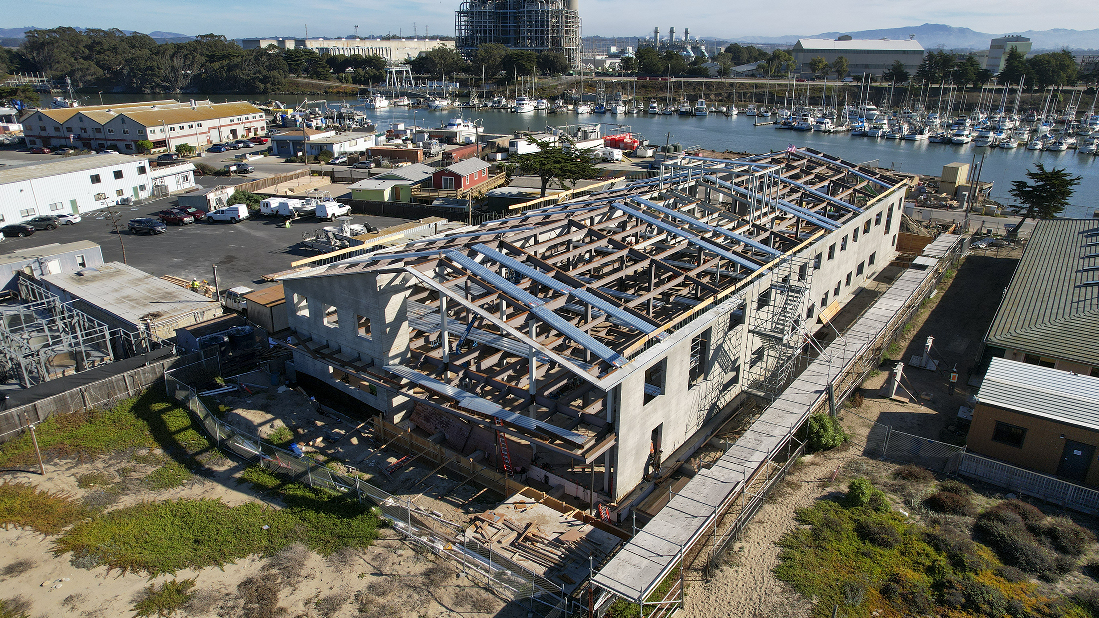 Aerial photo of construction of MBARI’s new robotic technology lab. The building’s gray concrete walls and metal rafters are in place, with wood beams and other internal elements installed inside. The beach with brown sand and green plants is visible in the foreground, a parking lot and buildings are visible to the left, and the Moss Landing Harbor and power plant are visible in the background.