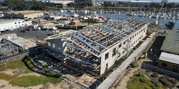 Aerial photo of construction of MBARI’s new robotic technology lab. The building’s gray concrete walls and metal rafters are in place, with wood beams and other internal elements installed inside. The beach with brown sand and green plants is visible in the foreground, a parking lot and buildings are visible to the left, and the Moss Landing Harbor and power plant are visible in the background.