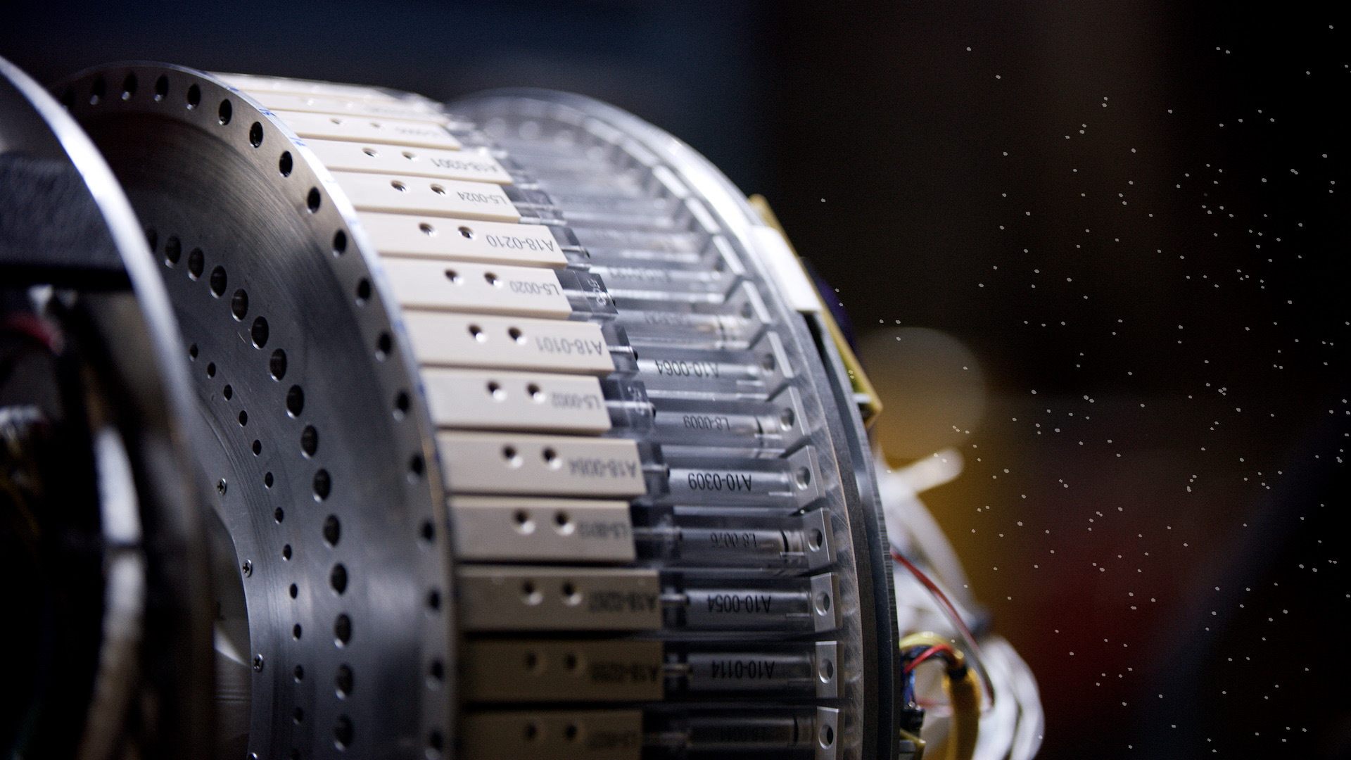 A carousel of beige plastic pucks with clear plastic plunger tops held on a circular metal plate. The pucks and plungers have alphanumeric codes printed on their sides in black ink. Metal instrumentation is on the left side of the frame, and multi-colored wires are on the right side of the frame. The background is blurred lens flare and animated white circular particles.