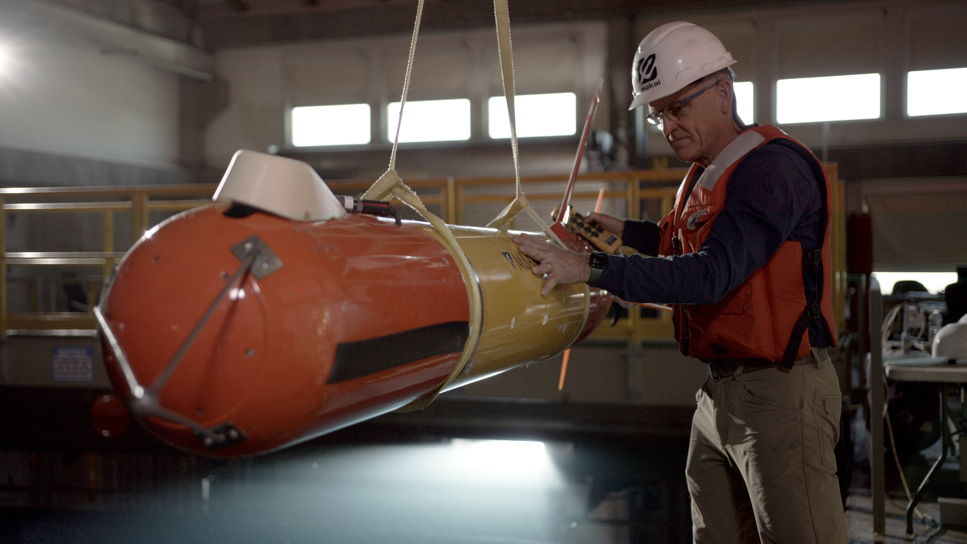 An MBARI engineer in an orange life vest, dark navy shirt, and brown pants wearing glasses and a white hard hat inspects an orange-and-yellow-colored, torpedo-shaped, autonomous robot. The engineer is holding a yellow control switch for an overhead crane. The robot is suspended from two yellow nylon straps. A large test tank is visible in the background.