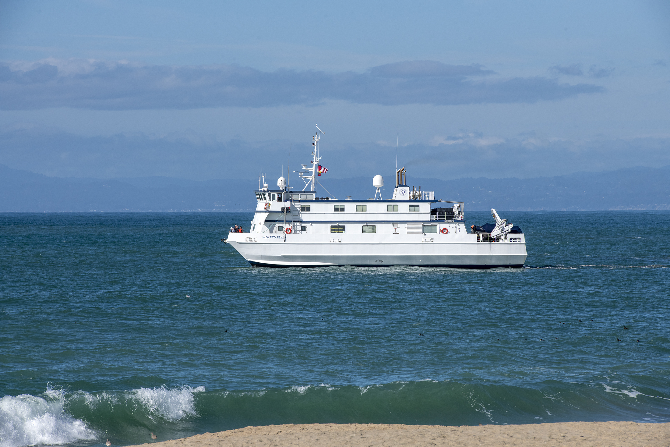 A white research vessel sailing in greenish-blue water with blue, cloudy sky visible in the background and green waves crashing on a sandy beach in the foreground.