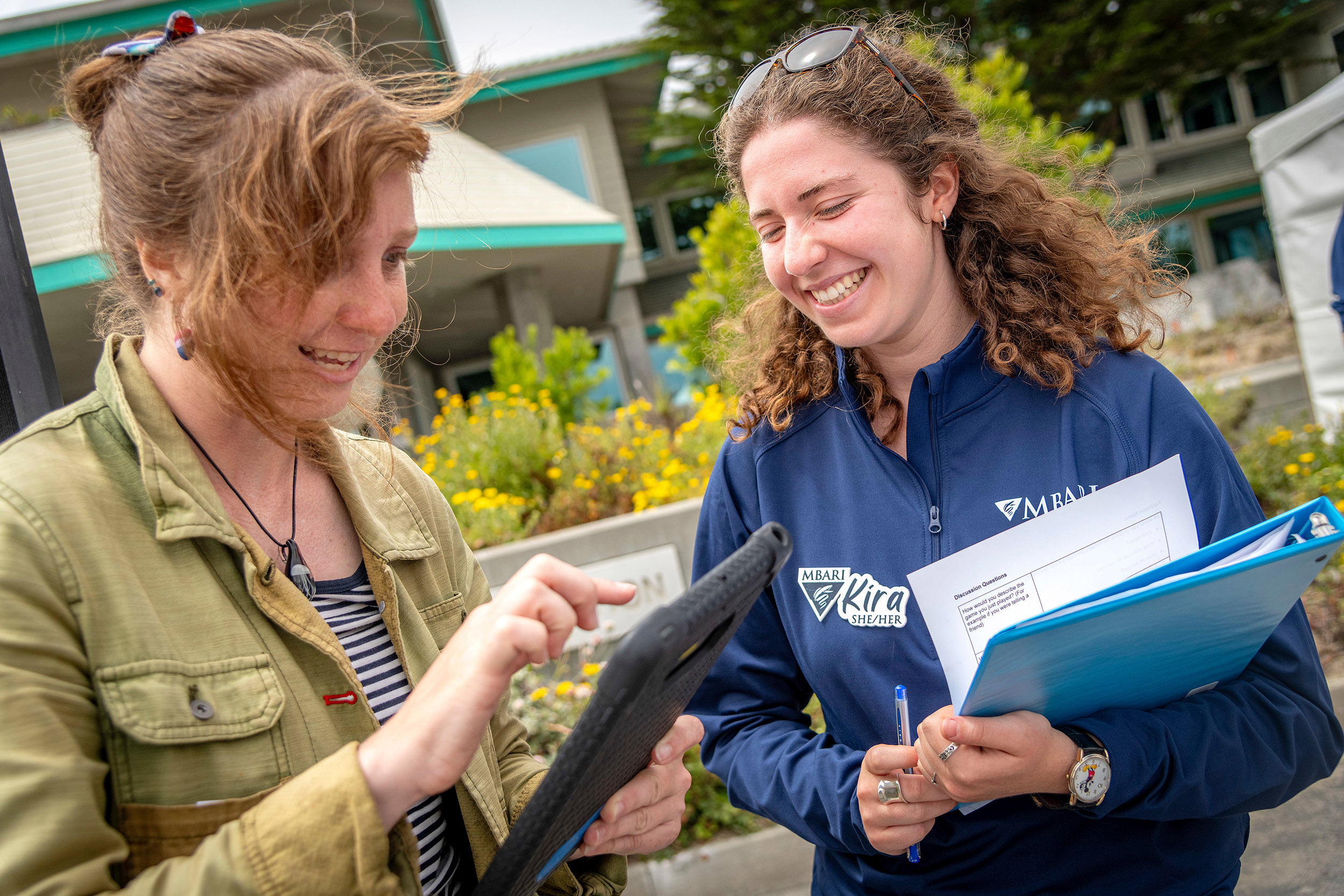An MBARI summer intern (right) with curly shoulder-length brown hair wearing a blue long-sleeved pullover and holding a blue binder and white papers speaks with an Open House visitor wearing a green jacket and black-and-white striped shirt and holding an iPad in a black rubberized case. This photograph was taken outdoors with MBARI buildings and green plants visible in the background.