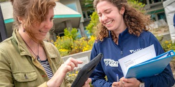 An MBARI summer intern (right) with curly shoulder-length brown hair wearing a blue long-sleeved pullover and holding a blue binder and white papers speaks with an Open House visitor wearing a green jacket and black-and-white striped shirt and holding an iPad in a black rubberized case. This photograph was taken outdoors with MBARI buildings and green plants visible in the background.