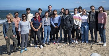 Sixteen interns assembled in a line posing for a photograph on the beach. Brown sand is visible in the foreground and greenish-blue ocean, white waves, and blue sky are visible in the background.