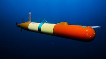 An orange-and-yellow-colored, torpedo-shaped autonomous robot gliding underwater. This screen capture from underwater video shows a robot against open blue water.