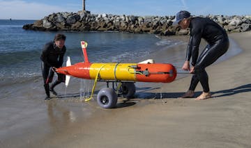 Two MBARI engineers in black wetsuits recover an orange-and-yellow-colored, torpedo-shaped autonomous robot using a black cart with large, gray wheels on the wet, sandy beach with a rocky jetty in the background.