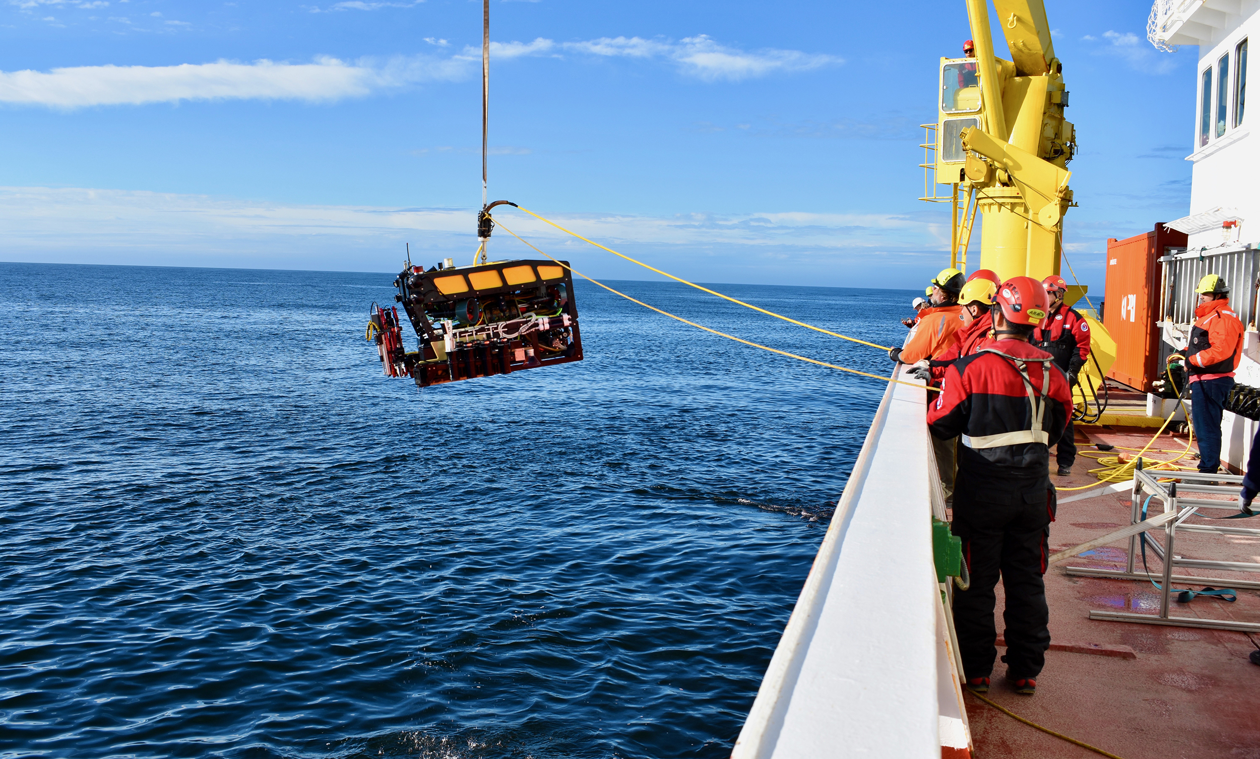 Marine operations crew members launch a robotic submersible into the water. The crew members are on the right side of the frame, lowering the submersible over the side of a research ship. The submersible has a yellow float, black frame, and a red mechanical arm, and is equipped with various scientific instruments. A black tether extending offscreen at the top of the frame is connected to a yellow crane on the research ship to lift the submersible from the ship into the water. Marine operations crew members are holding two yellow tethers, one to carry video from the submersible’s cameras and one to stabilize the submersible during deployment. The background is blue ocean and bright-blue sky with scattered streaks of white clouds.
