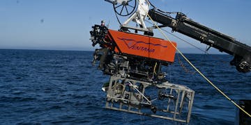 An MBARI remotely operated vehicle outfitted with a silver seafloor mapping toolsled. The robotic submersible has a bright orange float with the name “Ventana” with MBARI’s gulper eel logo serving as the “V.” Beneath the float is a black metal frame and various pieces of equipment and wires. The toolsled has a silver frame that extends past the back of the submersible. The submersible was photographed during recovery, attached to a black-and-white crane and positioned over the side of a research ship with dark blue water below and clear blue sky in the background. Four members of the ship crew are standing on the gray deck of the ship. Two are wearing bright orange life vests and white hard hats while managing the bright yellow tether connected to the submersible. One is wearing a bright orange jacket and spooling the tether. The fourth is wearing a bright orange jacket and holding a portable set of submersible controls.